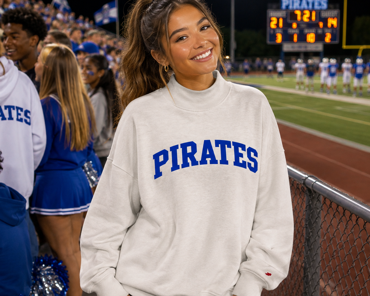 A smiling woman stands on a football field at night wearing a light gray sweatshirt with 'PIRATES' printed in blue across the chest. Cheerleaders and stadium crowds are visible in the background.