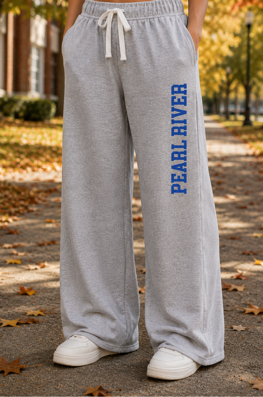 A person standing outdoors on a leaf-strewn path wearing light heather gray wide-leg sweatpants with white drawstrings and blue vertical 'PEARL RIVER' text on the left leg, paired with white sneakers.